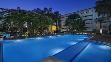 A large indoor swimming pool with clear lanes, surrounded by lounge chairs and palm trees, with a modern building in the background.