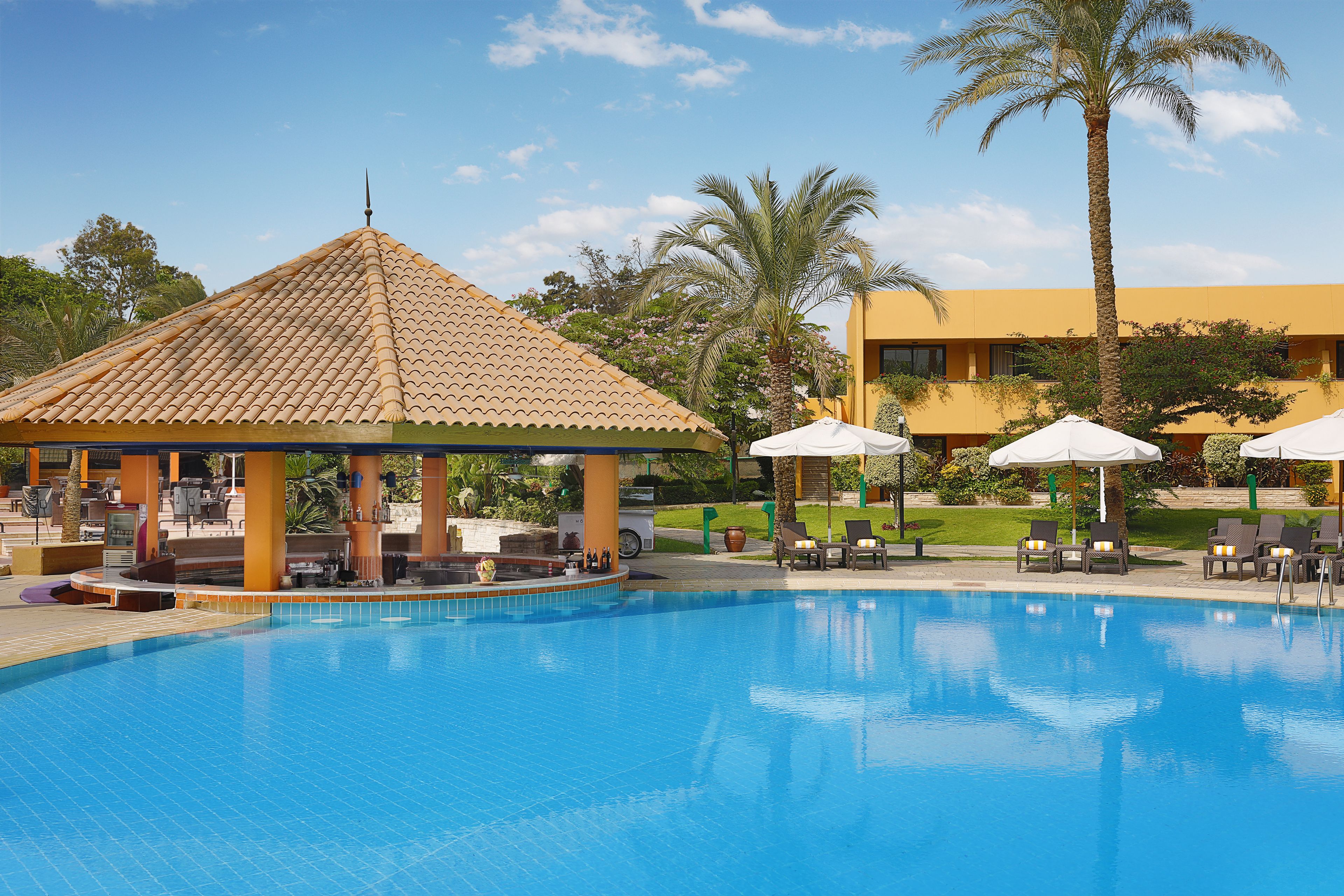 Ein Swimmingpool mit Liegestühlen und Sonnenschirmen, umgeben von Palmen und einem Gebäude mit einem Ziegeldach.