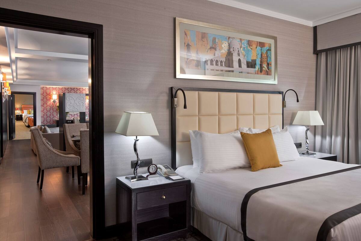 Quarto de hotel com uma cama grande, duas mesas de cabeceira, uma cadeira e uma escrivaninha com abajur.
