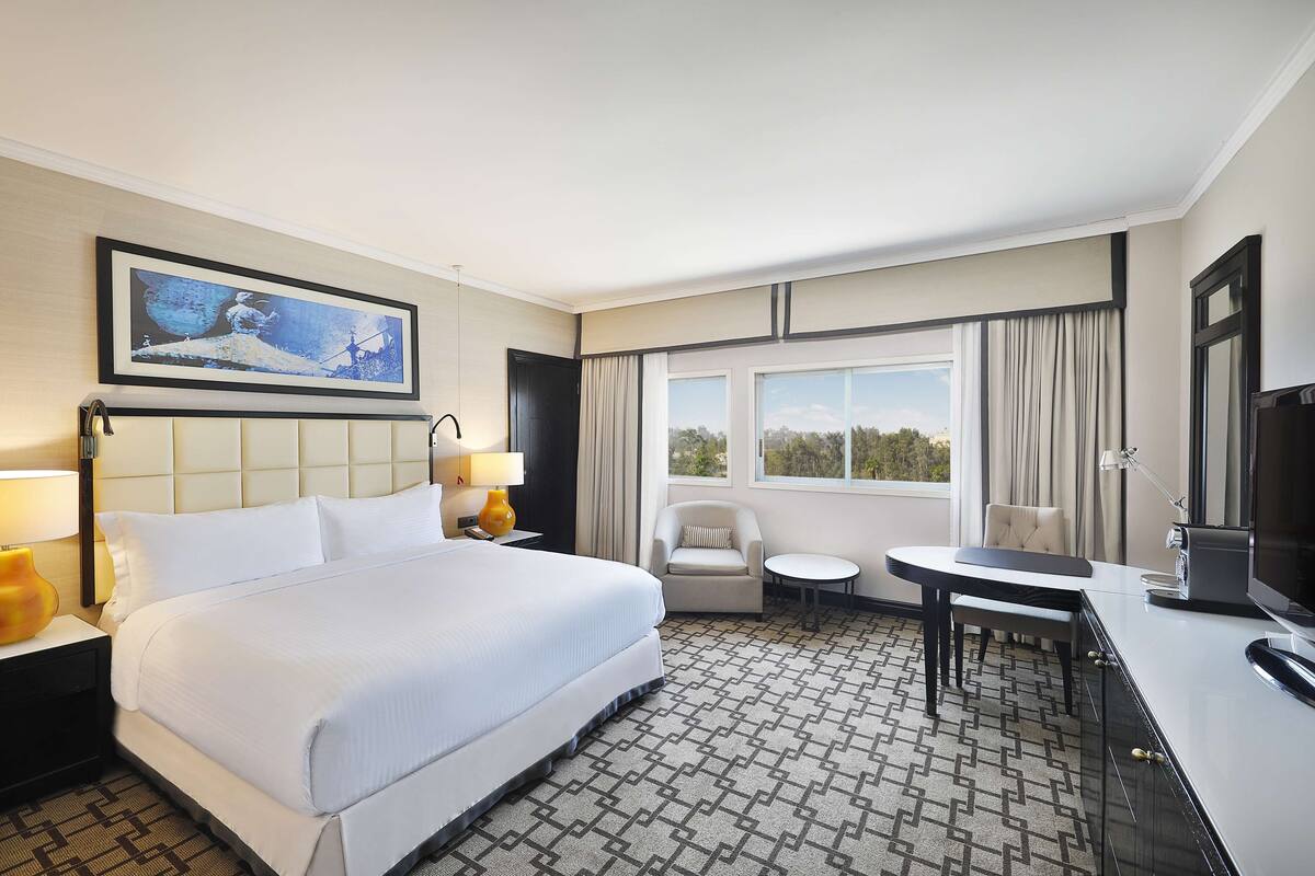 Quarto de hotel com uma cama grande, uma escrivaninha com cadeira, televisão e janela com cortinas.