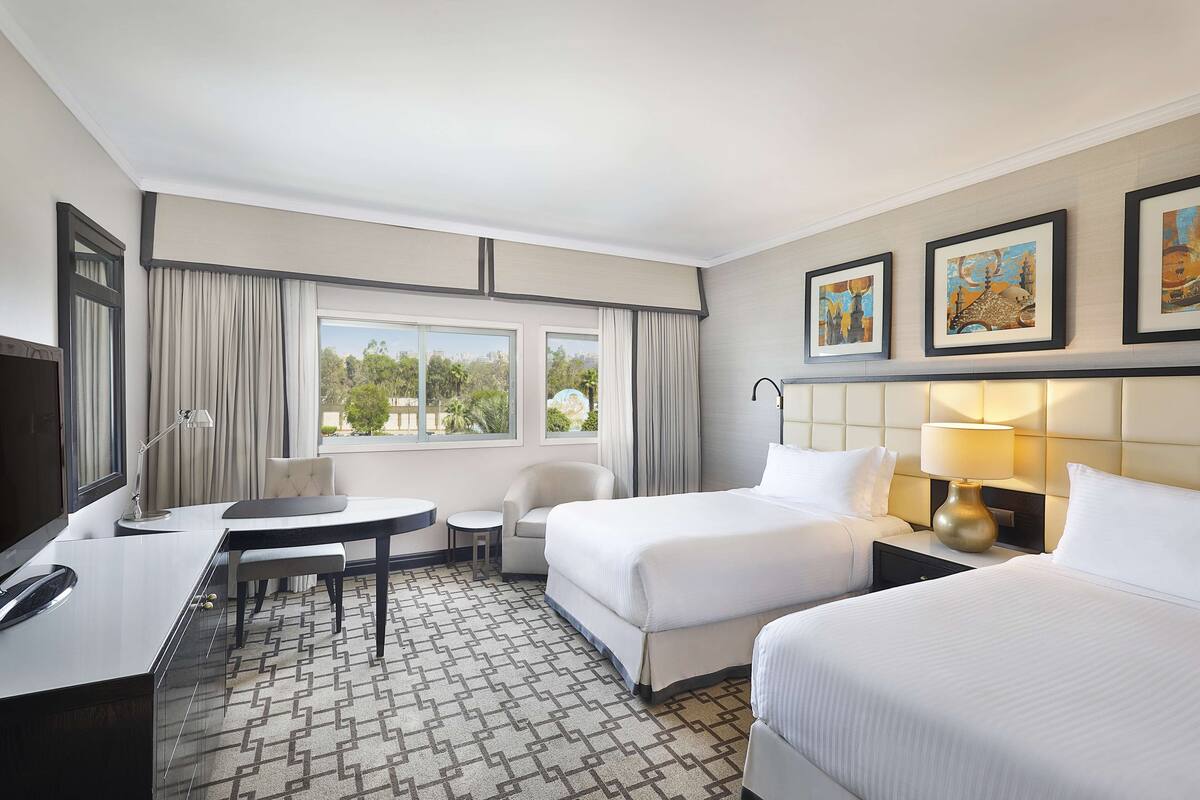 Quarto de hotel com duas camas, uma escrivaninha, uma cadeira, uma televisão e uma janela com cortinas.