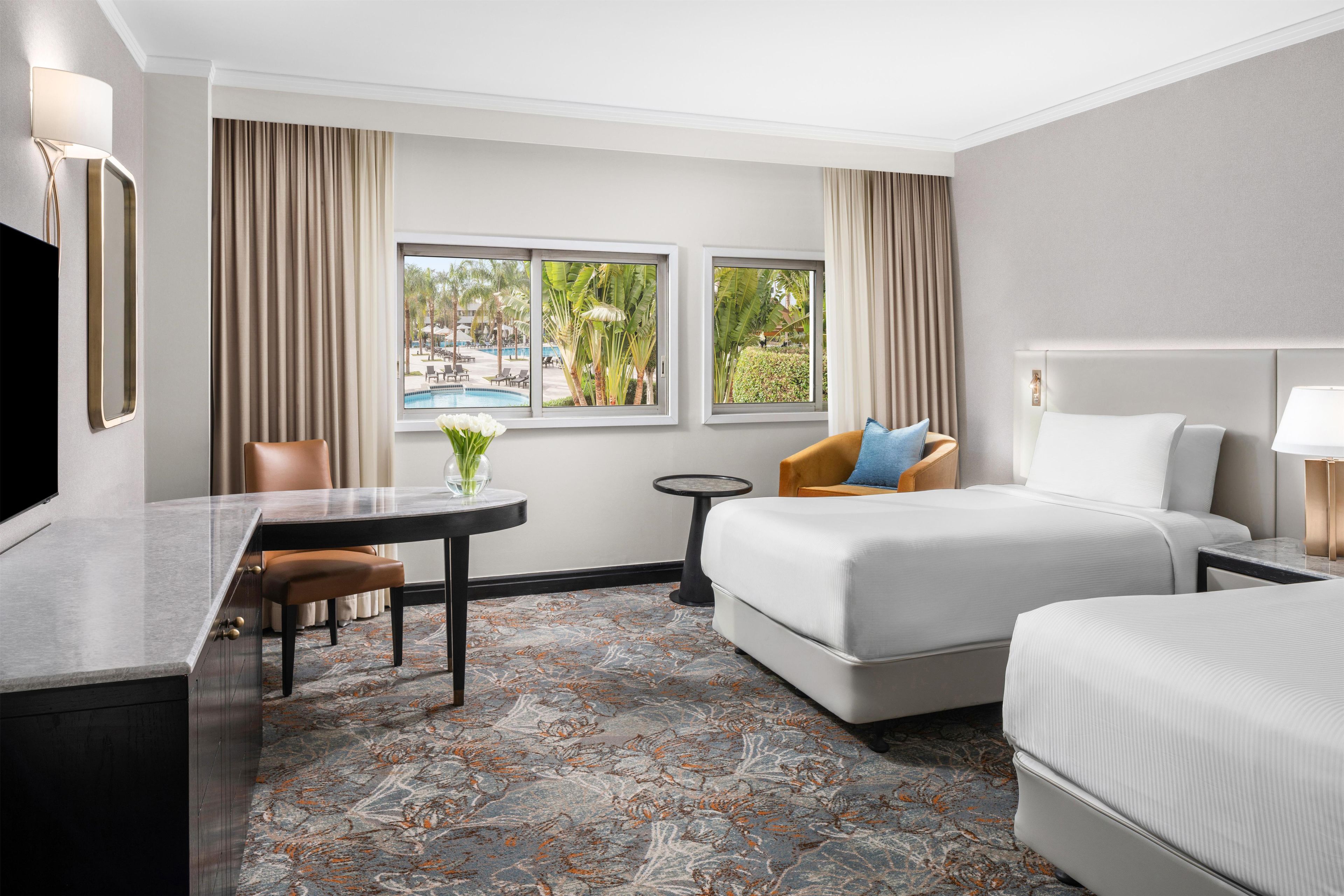 A hotel room with a bed, a desk, a chair, a small table, and a view of a pool.