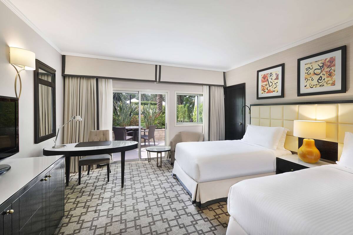 Quarto de hotel com duas camas, uma escrivaninha com cadeira, televisão e janela com cortinas.