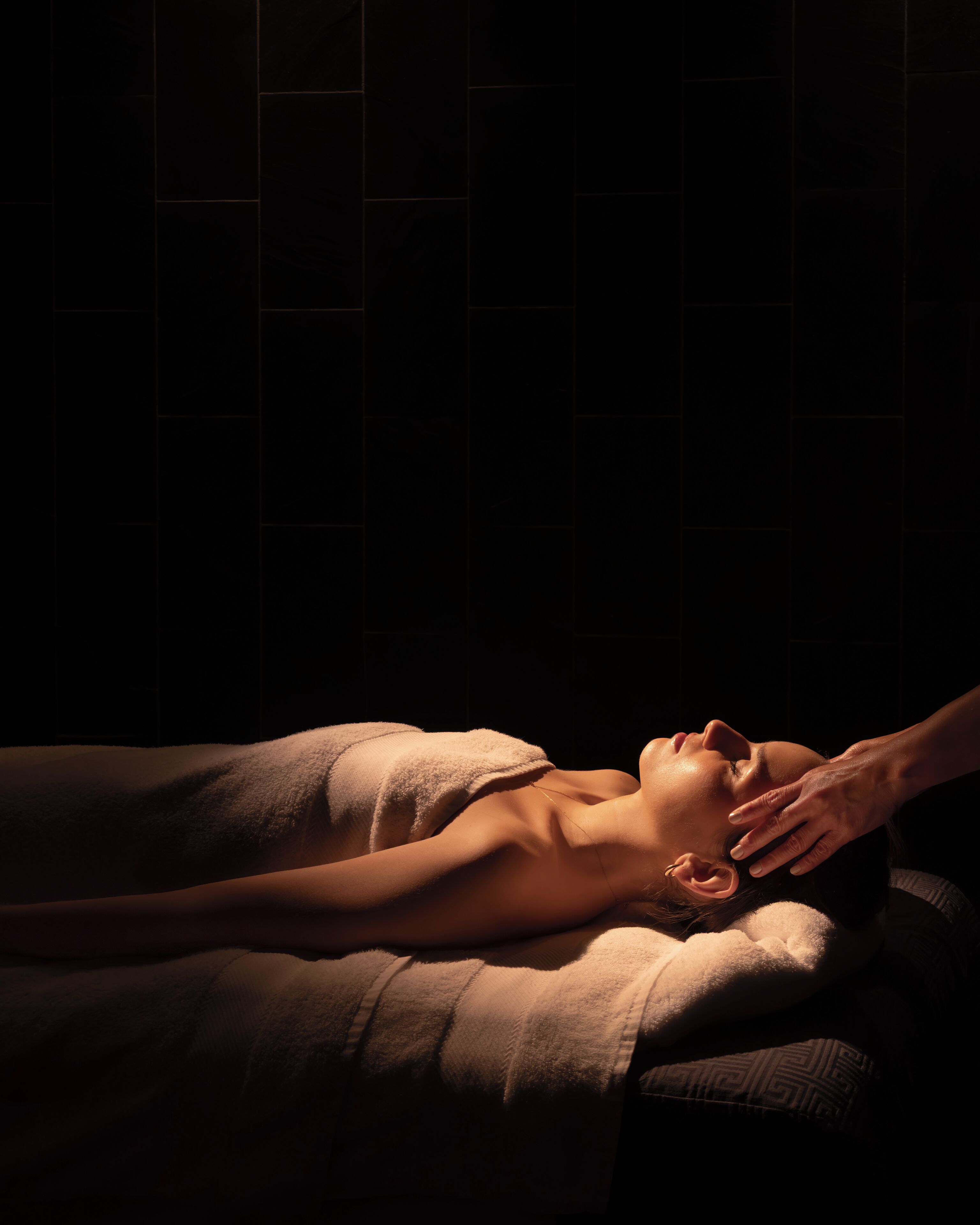 A person lying on a bed with a towel, in a dimly lit room with tiled walls.