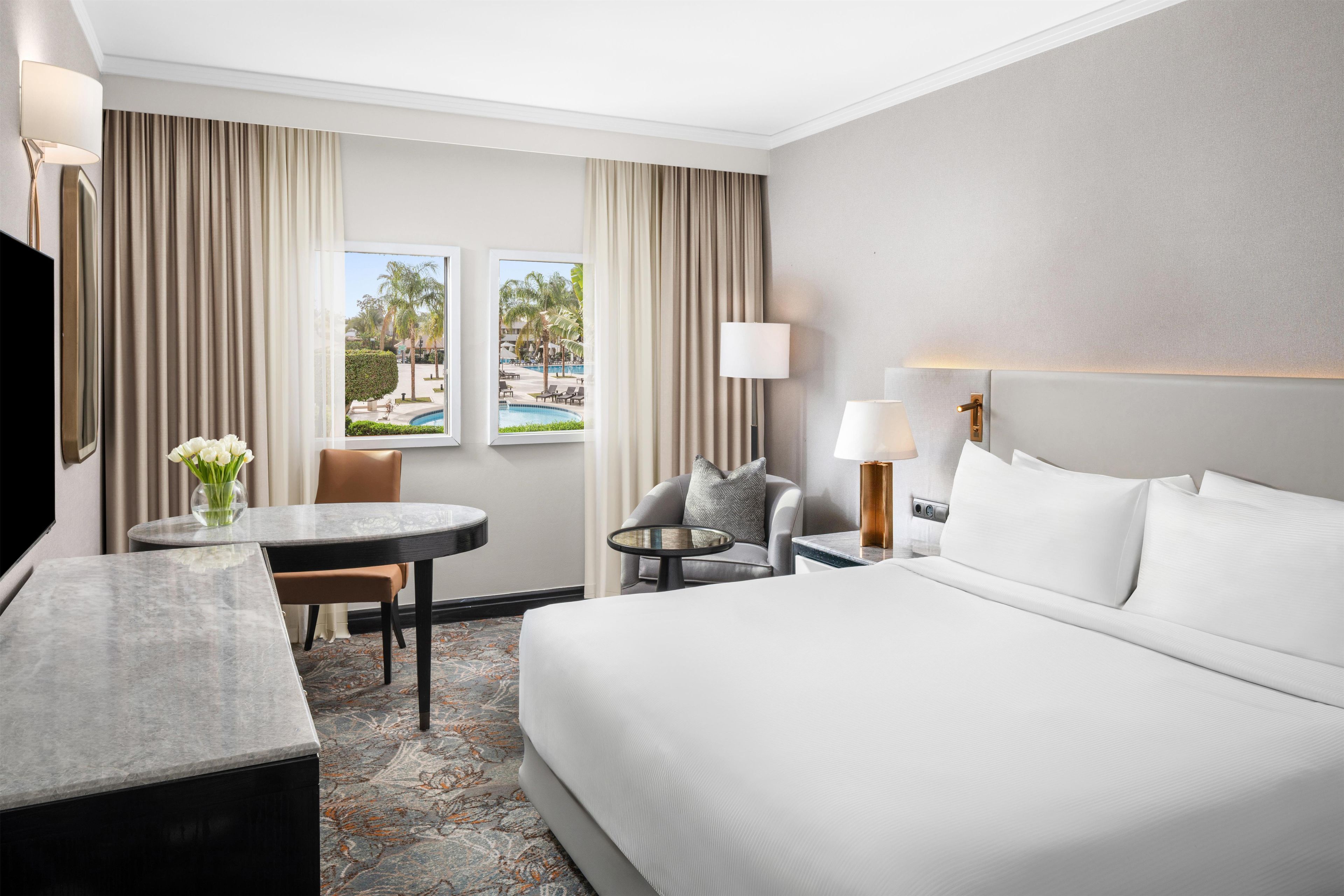 A hotel room with a bed, a desk, a chair, a TV, and a window with a view of a pool.