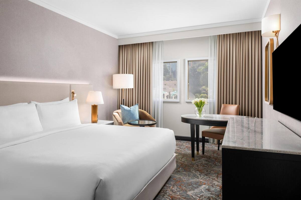 Quarto de hotel com uma cama grande, uma escrivaninha, uma cadeira, um abajur e uma televisão.