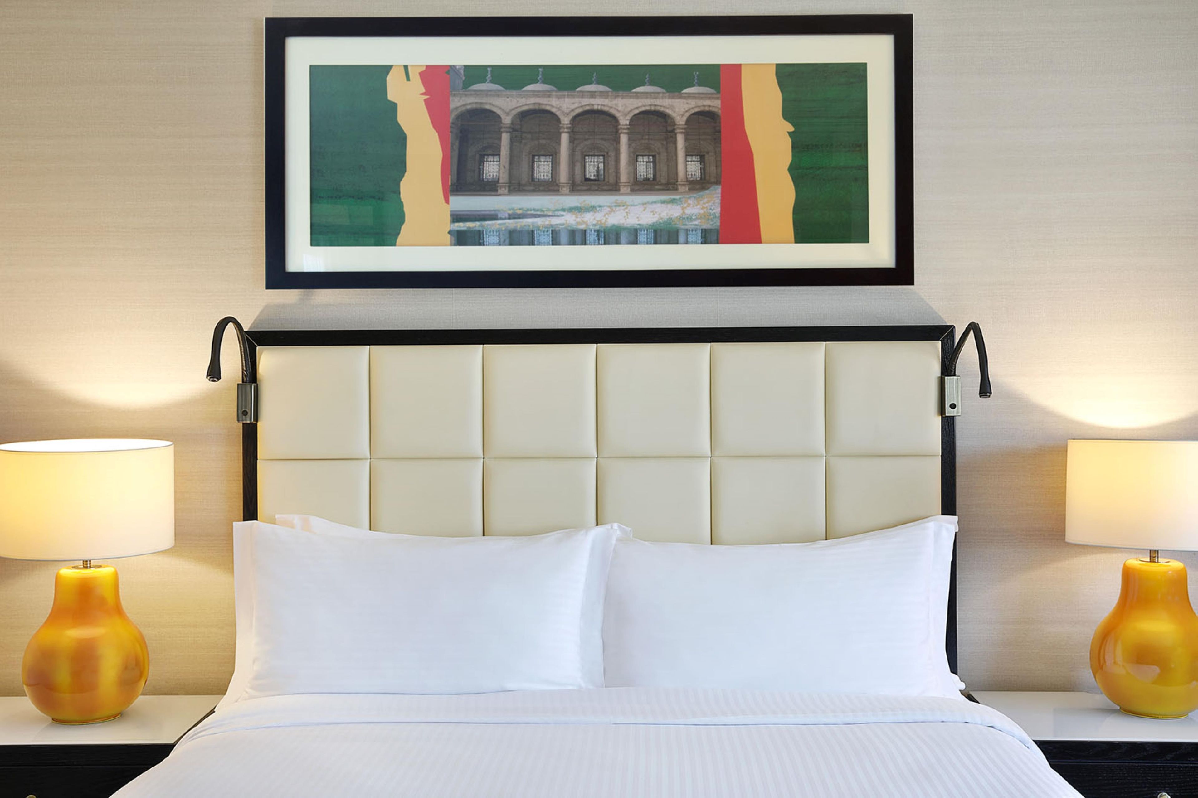 A hotel room with a bed, two bedside lamps, a framed artwork above the bed, and a headboard with a tufted design.