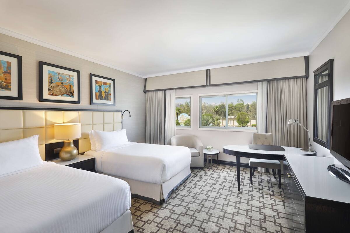 Quarto de hotel com duas camas, uma mesa, uma cadeira, uma televisão e uma janela com cortinas.