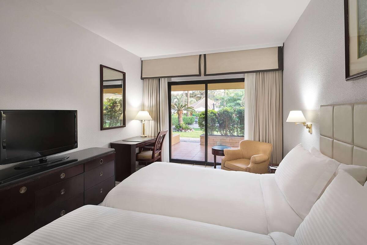 Quarto de hotel com cama, televisão, escrivaninha e cadeira. Porta de vidro deslizante dá acesso a área externa com plantas e vista do céu.