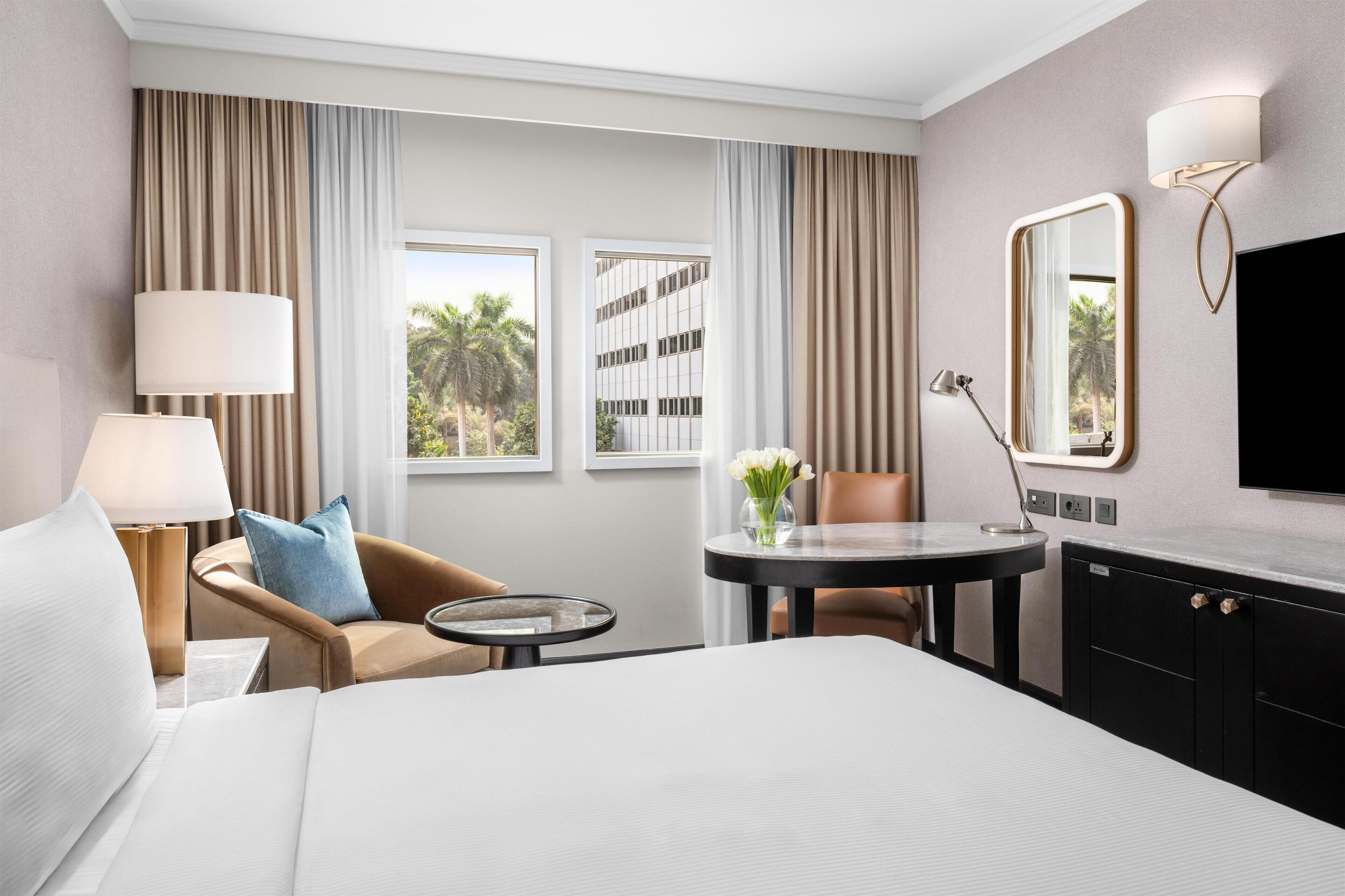 Habitación de hotel con cama, zona de estar con una silla y una mesa, televisor de pantalla plana, lámpara y ventana con cortinas.