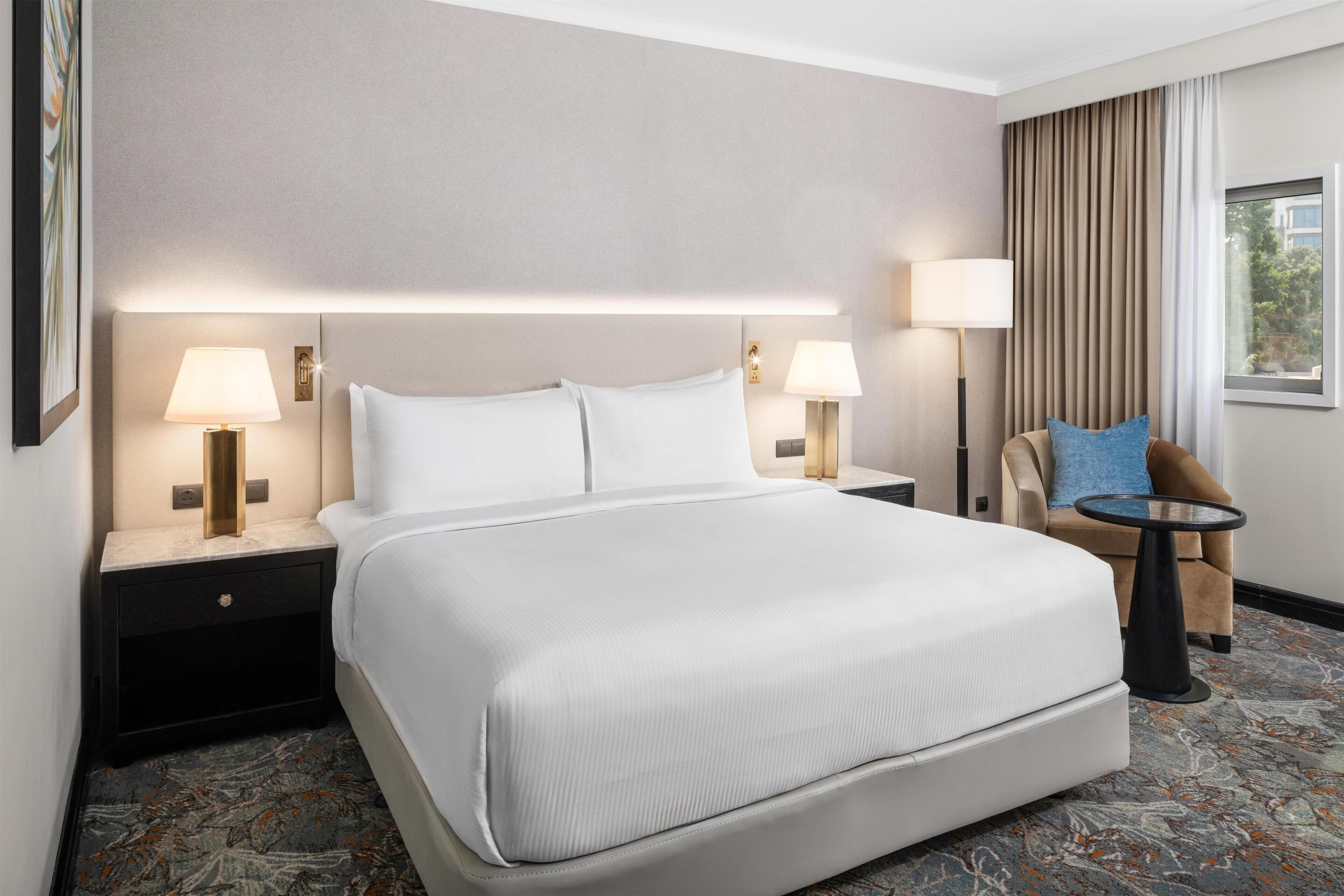 Habitación de hotel con una cama grande, dos mesitas de noche con lámparas, una silla y una pequeña mesa.