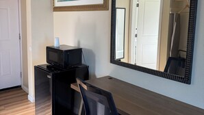 A room with a wooden desk, a black office chair, a black refrigerator, a microwave, a framed painting, and a large mirror. - Castaway Motel (Orange)