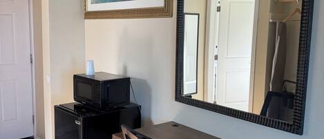 Quarto com escrivaninha de madeira, cadeira de escritório preta, geladeira preta, micro-ondas, quadro emoldurado e um espelho grande.