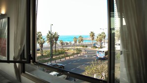 A view from a window overlooking a street with palm trees, a bus, and a beach in the distance. - Panoramic Ocean View 1 min From the Sea (Tel Aviv)
