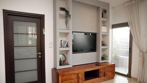 A modern living room with a large flat-screen TV mounted on a wooden console, a sliding glass door, and a bookshelf with decorative items. - Panoramic Ocean View 1 min From the Sea (Tel Aviv)