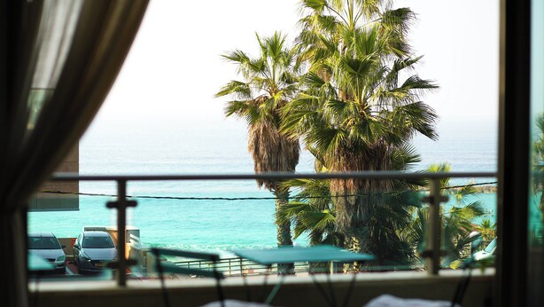 A balcony with a view of the ocean and palm trees. There are two cars parked outside and a table with chairs. - Panoramic Ocean View 1 min From the Sea (Tel Aviv)
