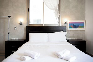 A neatly made bed with white linens and towels, a black nightstand with a lamp, and a painting on the wall. - Panoramic Ocean View 1 min From the Sea (Tel Aviv)
