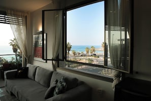 A living room with a large window overlooking a beach, a sofa, and a potted plant. - Panoramic Ocean View 1 min From the Sea (Tel Aviv)