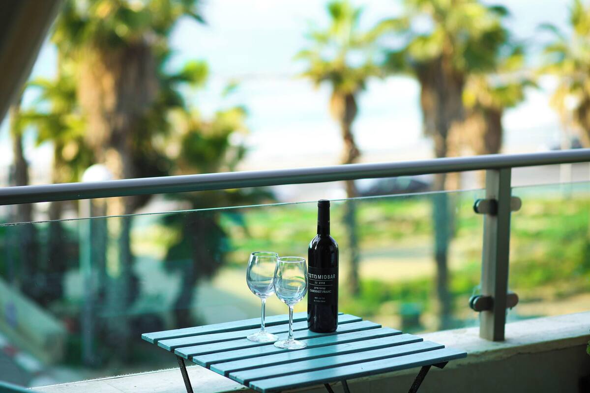 A table with two wine glasses and a bottle of wine on a balcony with a view of palm trees.