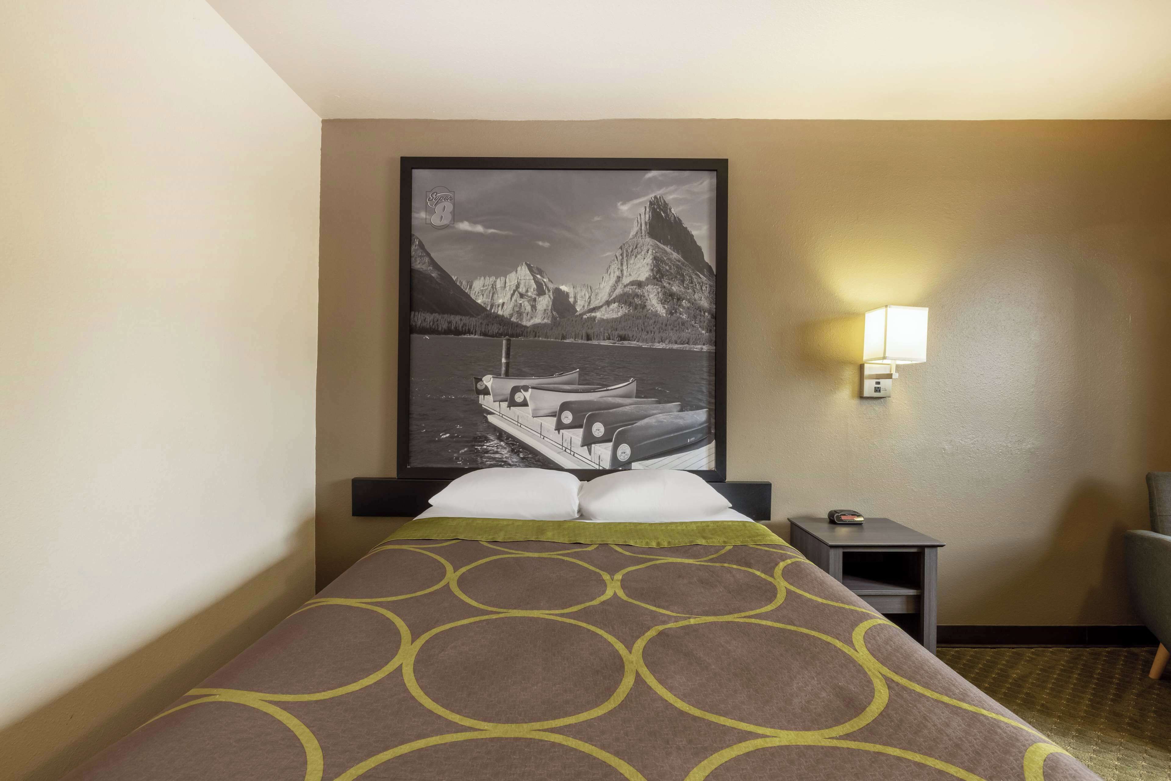 Habitación de hotel con una cama, una mesita de noche, una lámpara y un cuadro con un paisaje de montaña.