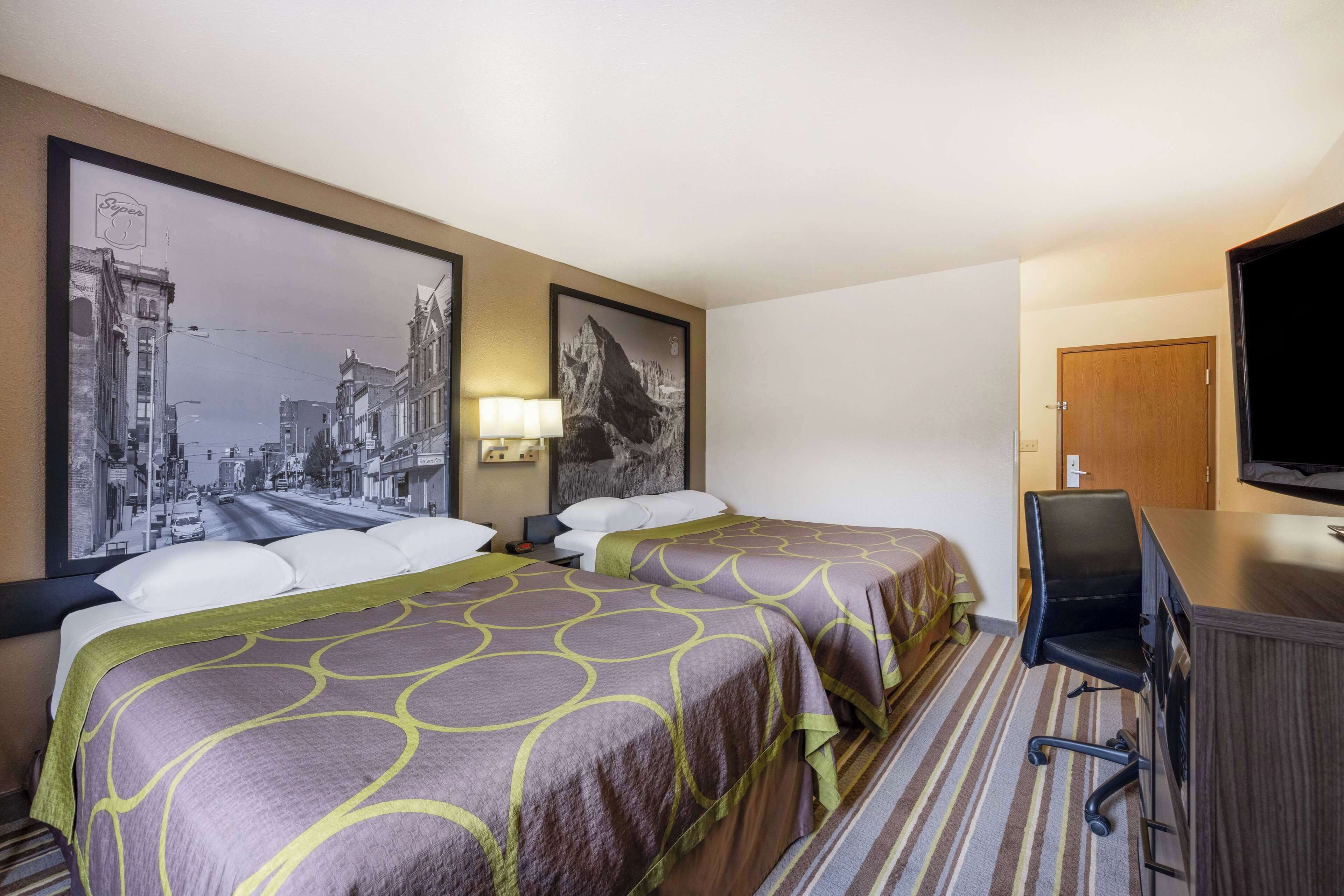 A hotel room with two beds, a desk, a chair, a television, and framed pictures on the wall.