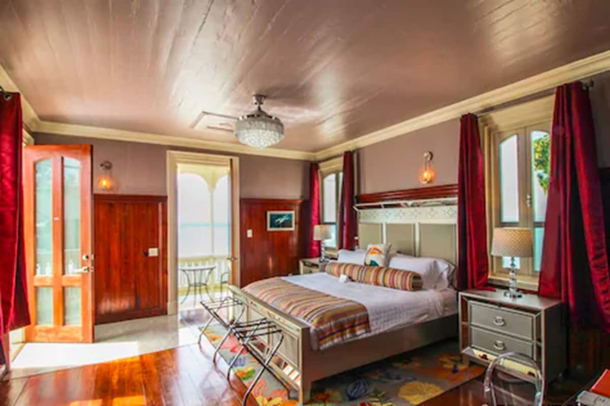 A bedroom with a wooden ceiling, a bed with a striped blanket, a nightstand, a lamp, and a window with red curtains.