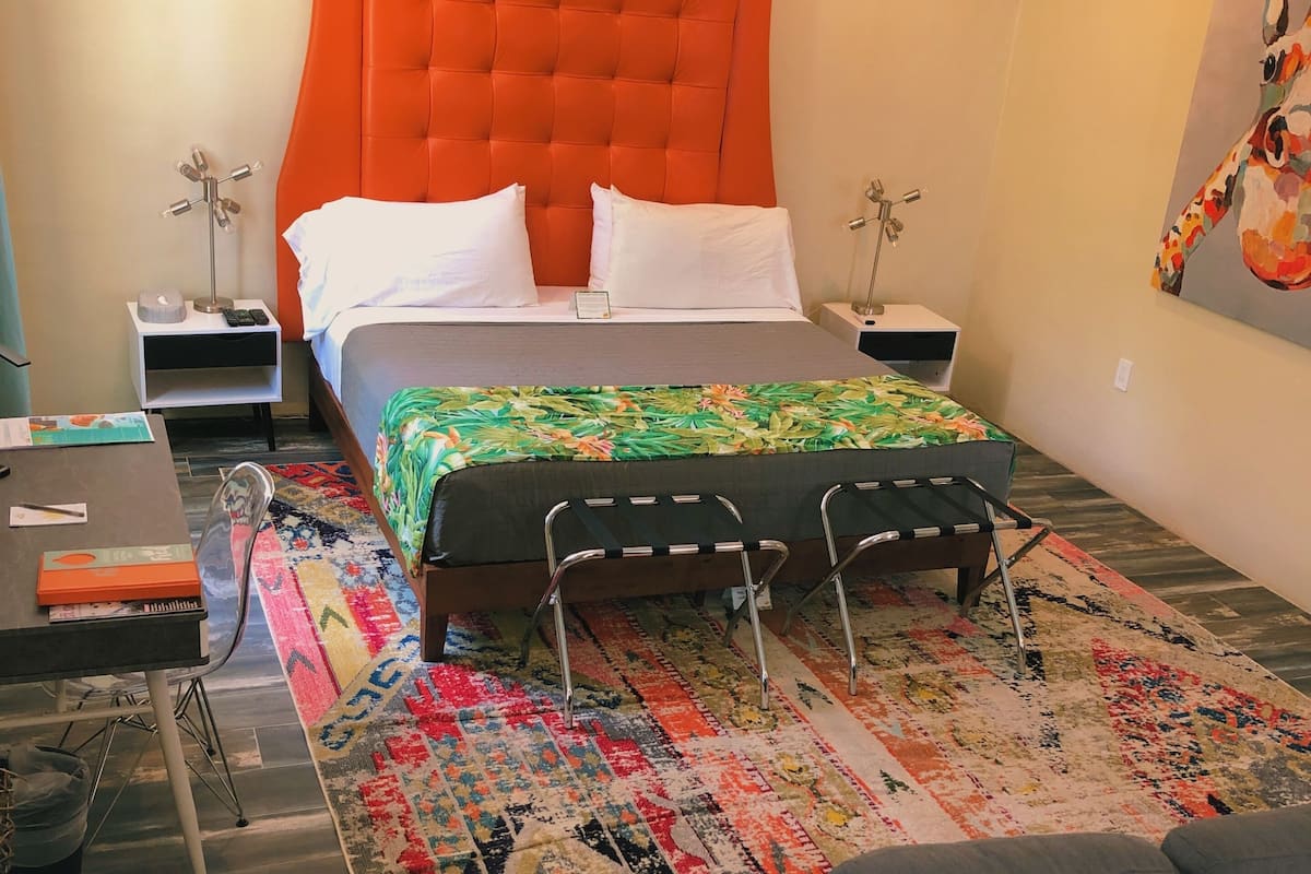 A hotel room with a large orange headboard, a bed with a colorful floral bedspread, a gray sofa, and a patterned rug.