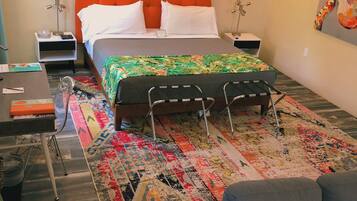 A hotel room with a large orange headboard, a bed with a colorful floral bedspread, a gray sofa, and a patterned rug.