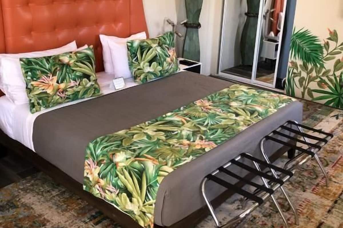 A bedroom with a tufted headboard, a bed with a tropical leaf-patterned bedspread, a small table with a chair, and a large mirror.