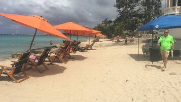 A beach with rows of lounge chairs and orange umbrellas, a man walking, and a building with a balcony in the background.