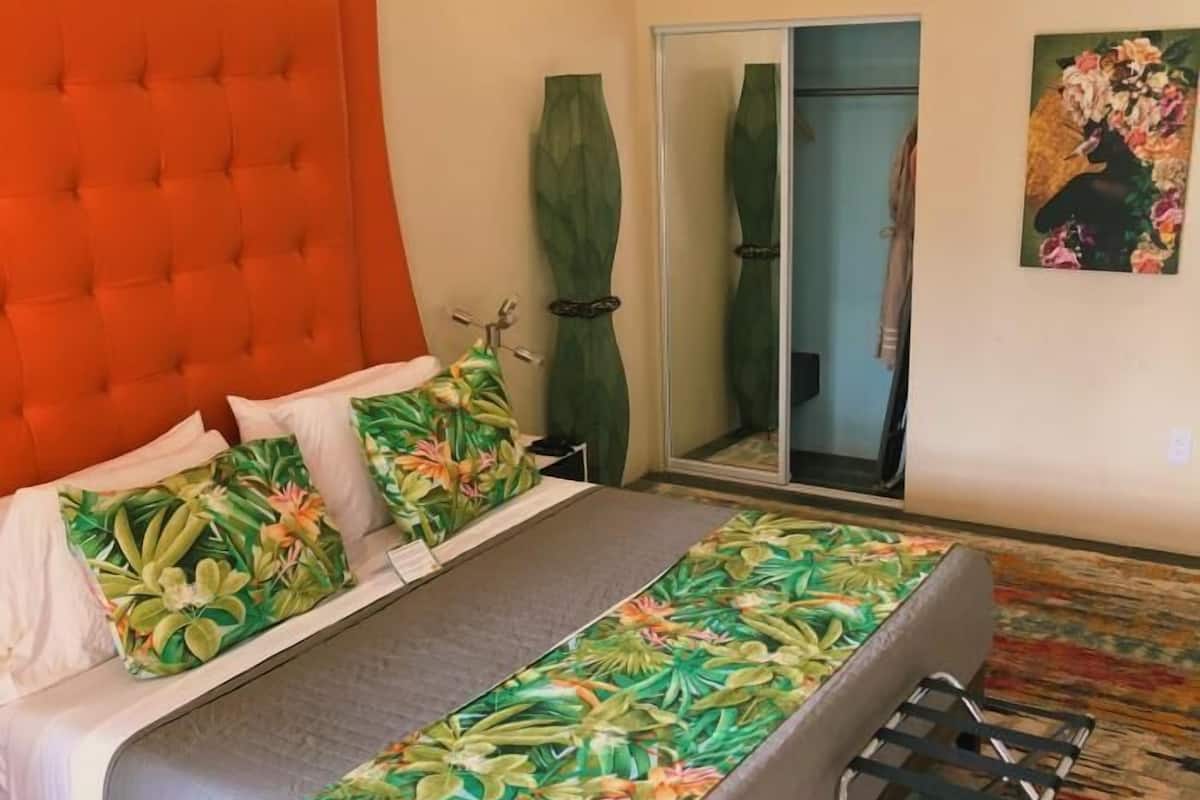 A bedroom with a tufted orange headboard, a bed with floral bedding, a green vase, and a colorful carpet.