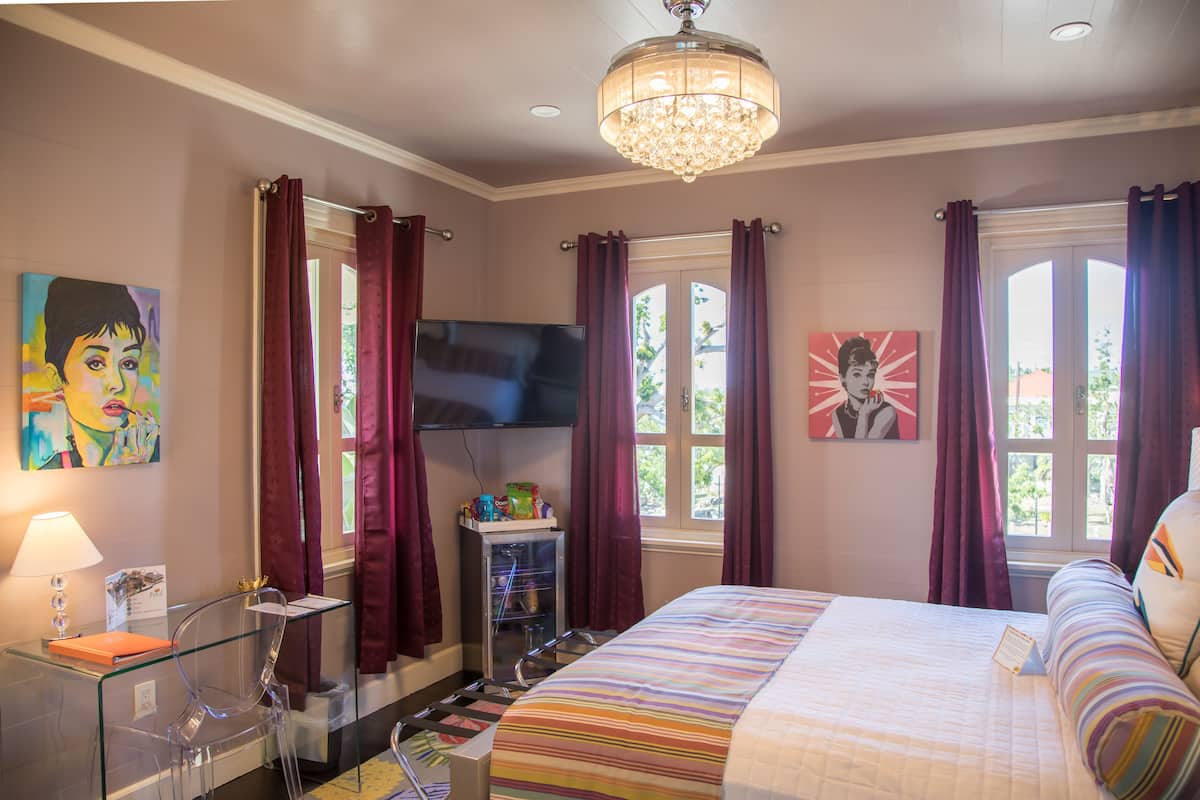 A bedroom with a bed, a TV, a desk with a lamp, and artwork on the walls.
