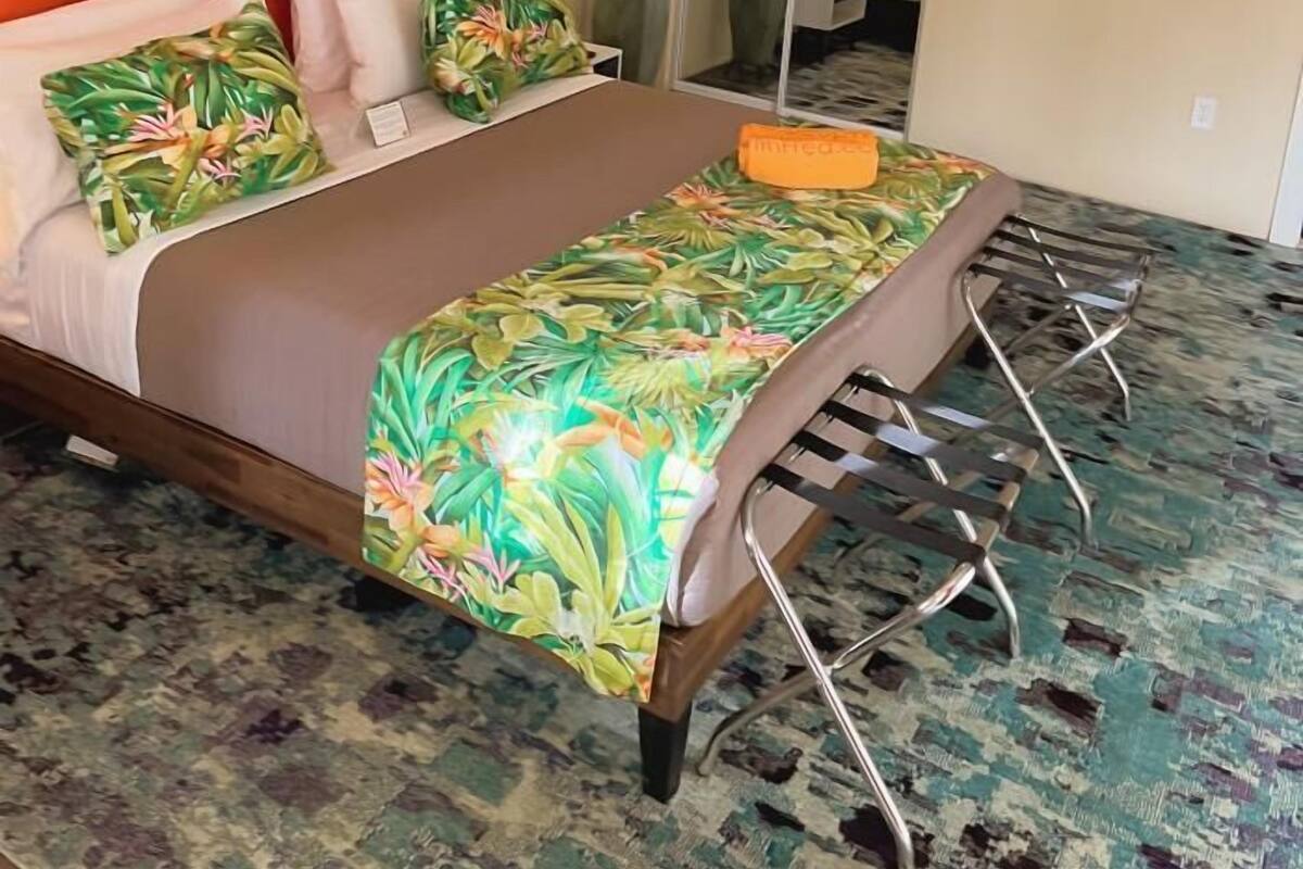 A bedroom with a large bed, an air conditioning unit, a ceiling fan, a mirror, and a colorful painting.
