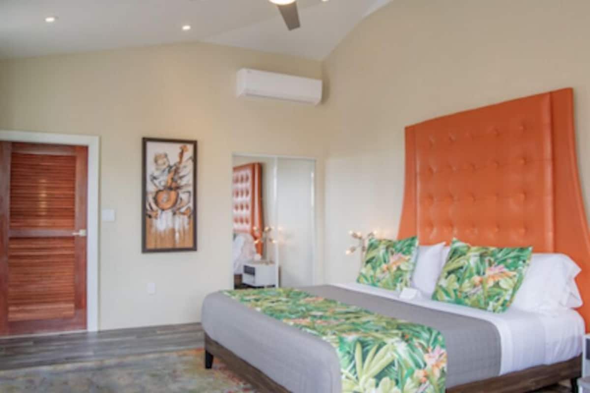 A bedroom with a large bed, a wooden door, a framed picture on the wall, and a ceiling fan.