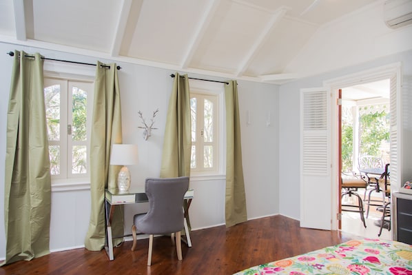 Una habitación con escritorio, silla y cama con colcha estampada. Hay dos ventanas con cortinas, una lámpara de pared y un adorno en la pared.