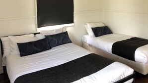 Dos camas individuales con ropa de cama blanca y negra, una mesita con un control remoto y una ventana con persianas.