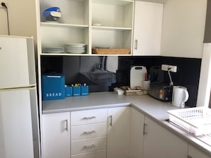 Una kitchenette compacta con armarios blancos, cafetera y tostadora. Hay cajas azules de almacenamiento con las etiquetas 'TÉ', 'AZÚCAR' y 'CAFÉ'.