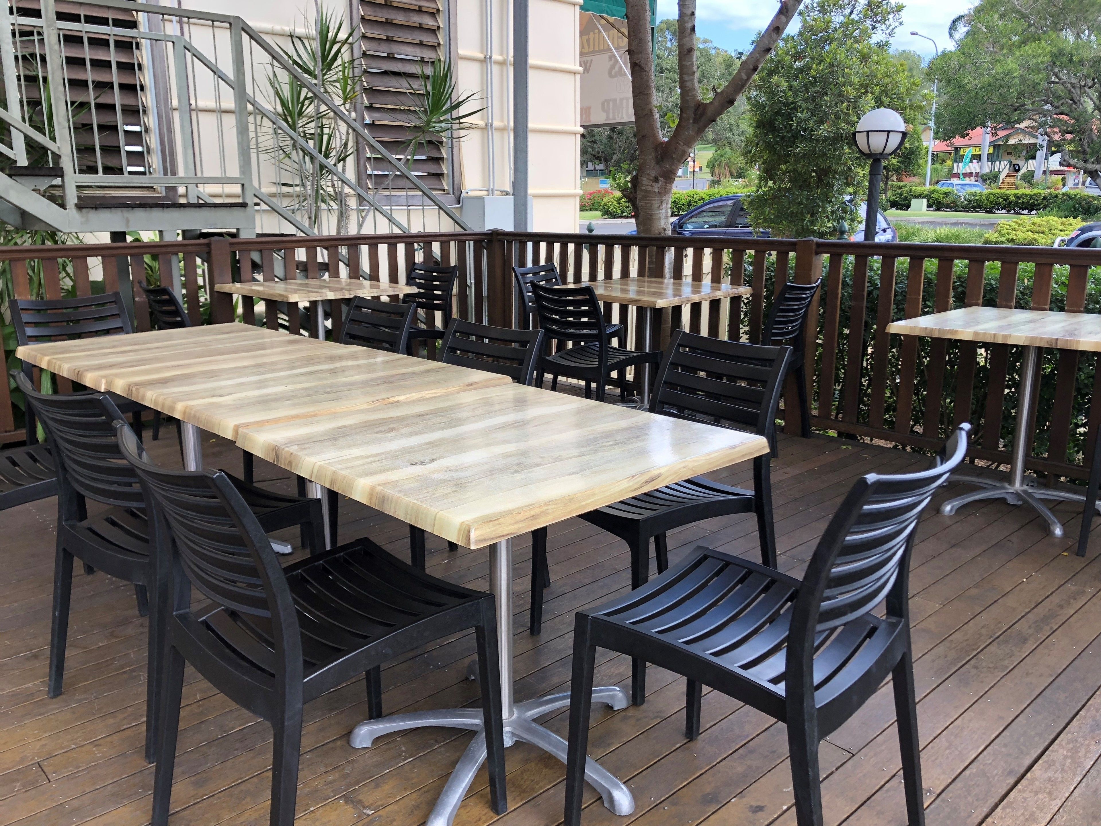 An outdoor patio with wooden tables and black chairs, surrounded by a wooden deck and a metal railing.
