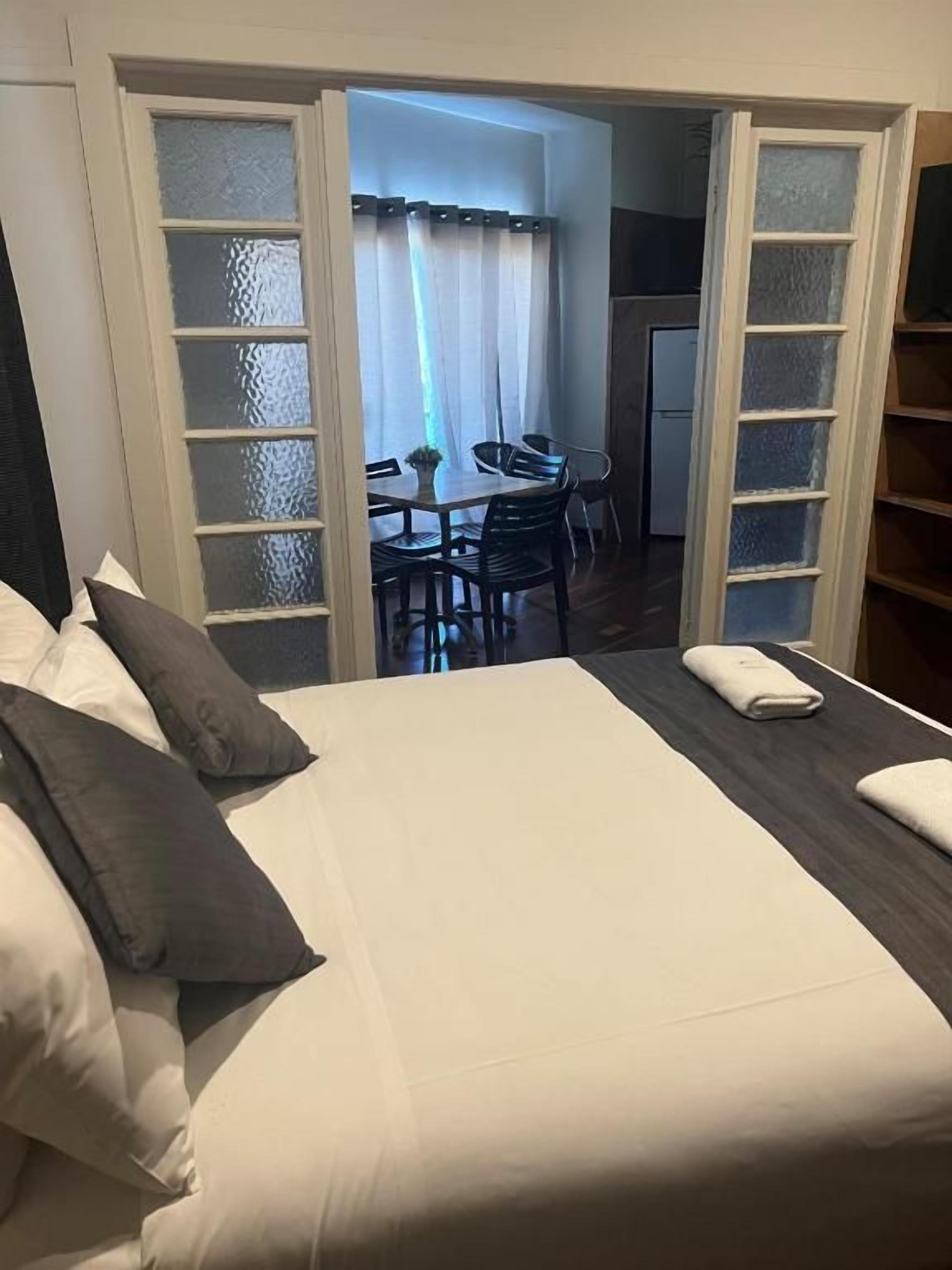 A hotel room with a bed, a small table, and a chair. There is a door leading to a dining area.