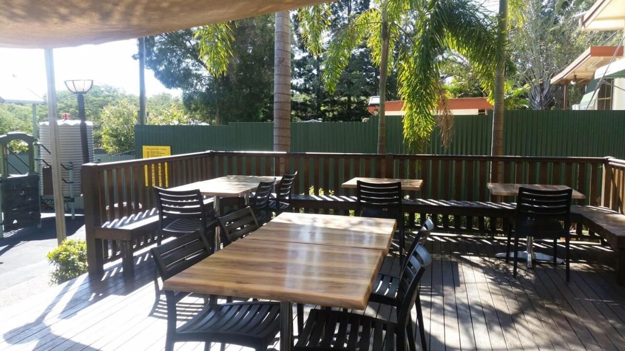 Een terras buiten met houten tafels en stoelen, omgeven door een groene hek en bomen.