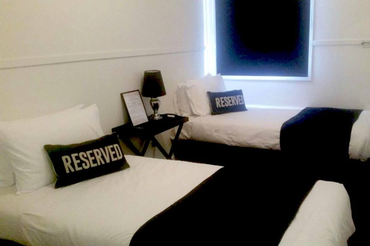 Habitación de hotel con dos camas, cada una con una almohada que dice 'RESERVED', una mesita de noche con una lámpara y una ventana con persianas.