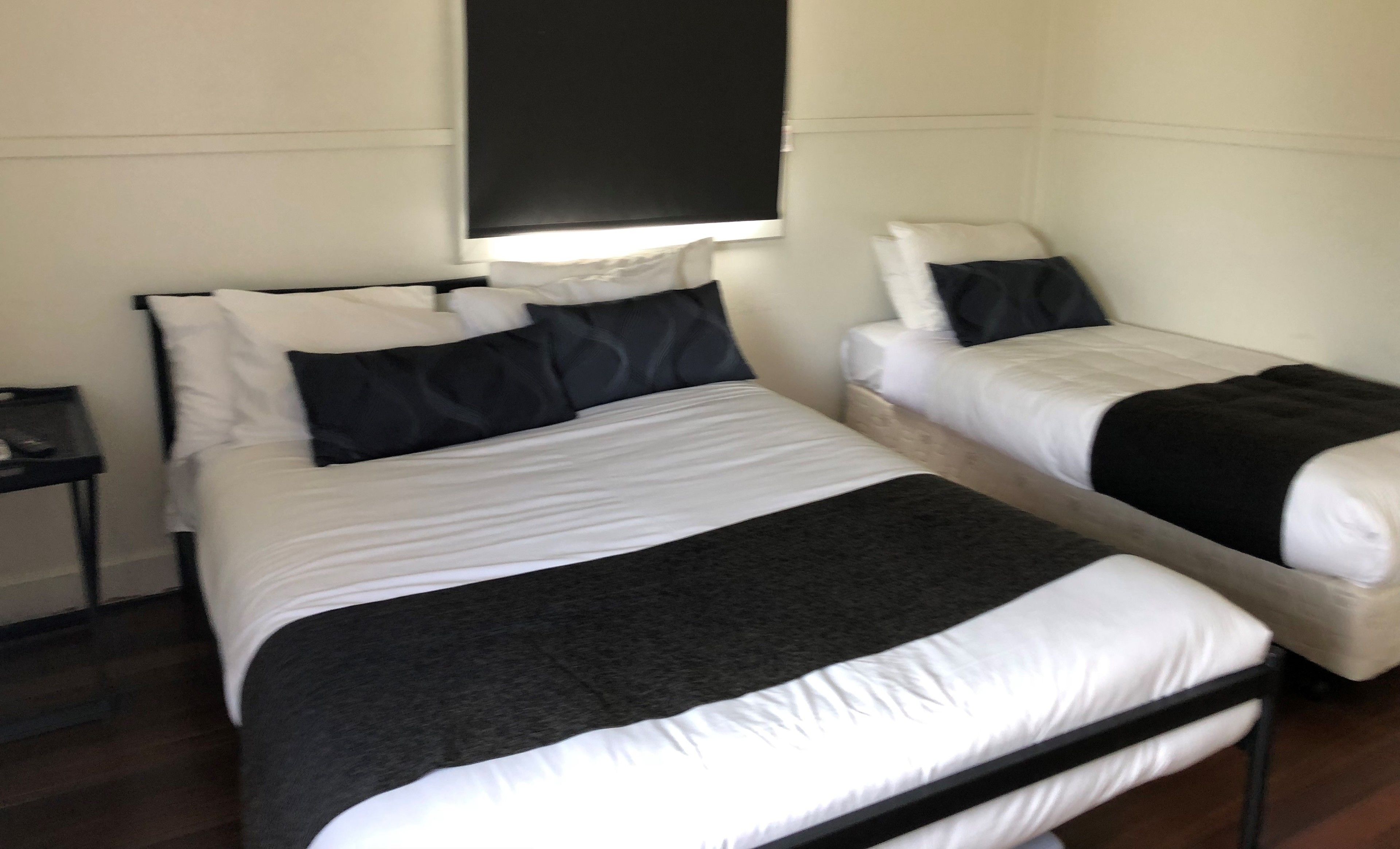 A room with two beds, each with white bedding and black headboards. There is a small table beside one bed and a wall-mounted shelf above the other bed.