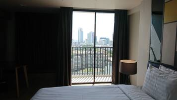 Ein Hotelzimmer mit Bett, Schreibtisch, Lampe und Blick auf eine Stadtlandschaft durch ein großes Fenster.