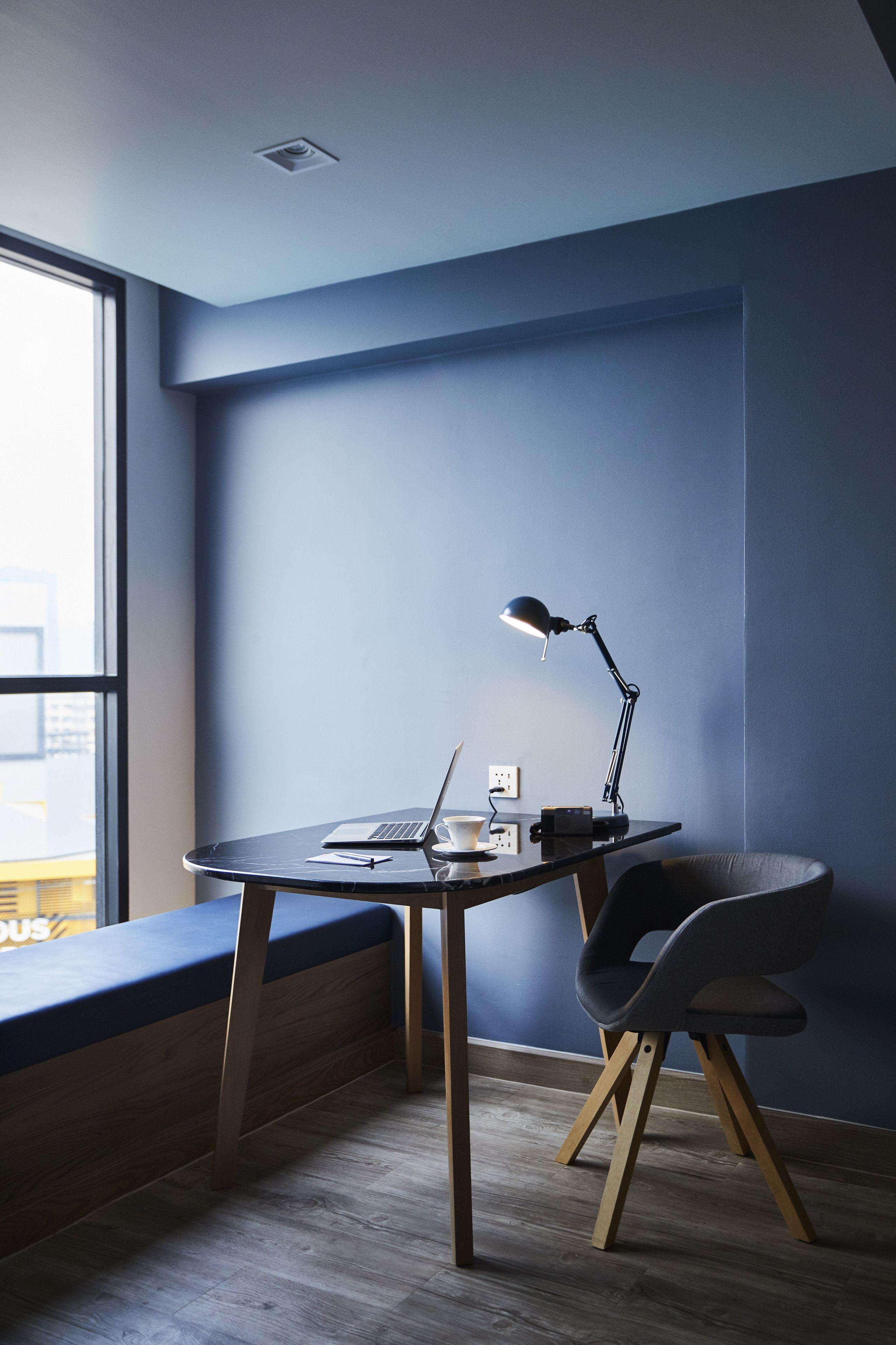 A modern workspace with a laptop, desk lamp, and chair, featuring a large window and a blue wall.