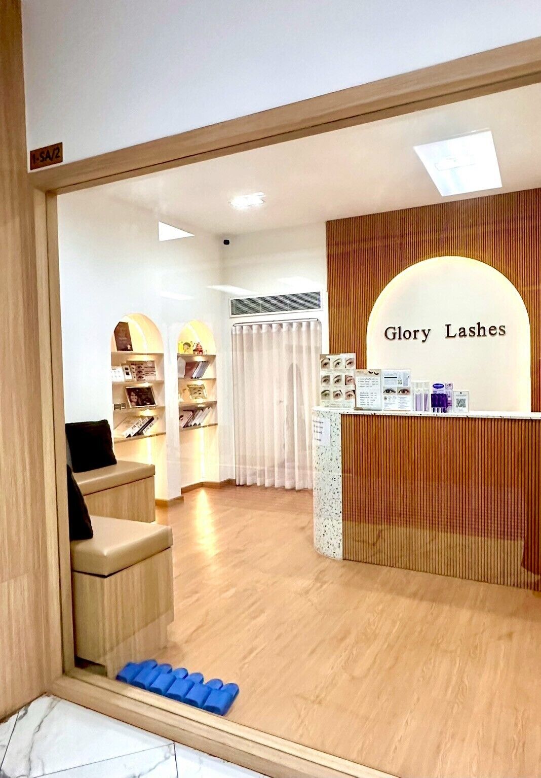 A salon interior with a reception desk labeled 'Glory Lashes,' a waiting area with a counter and shelves, and a wooden floor.