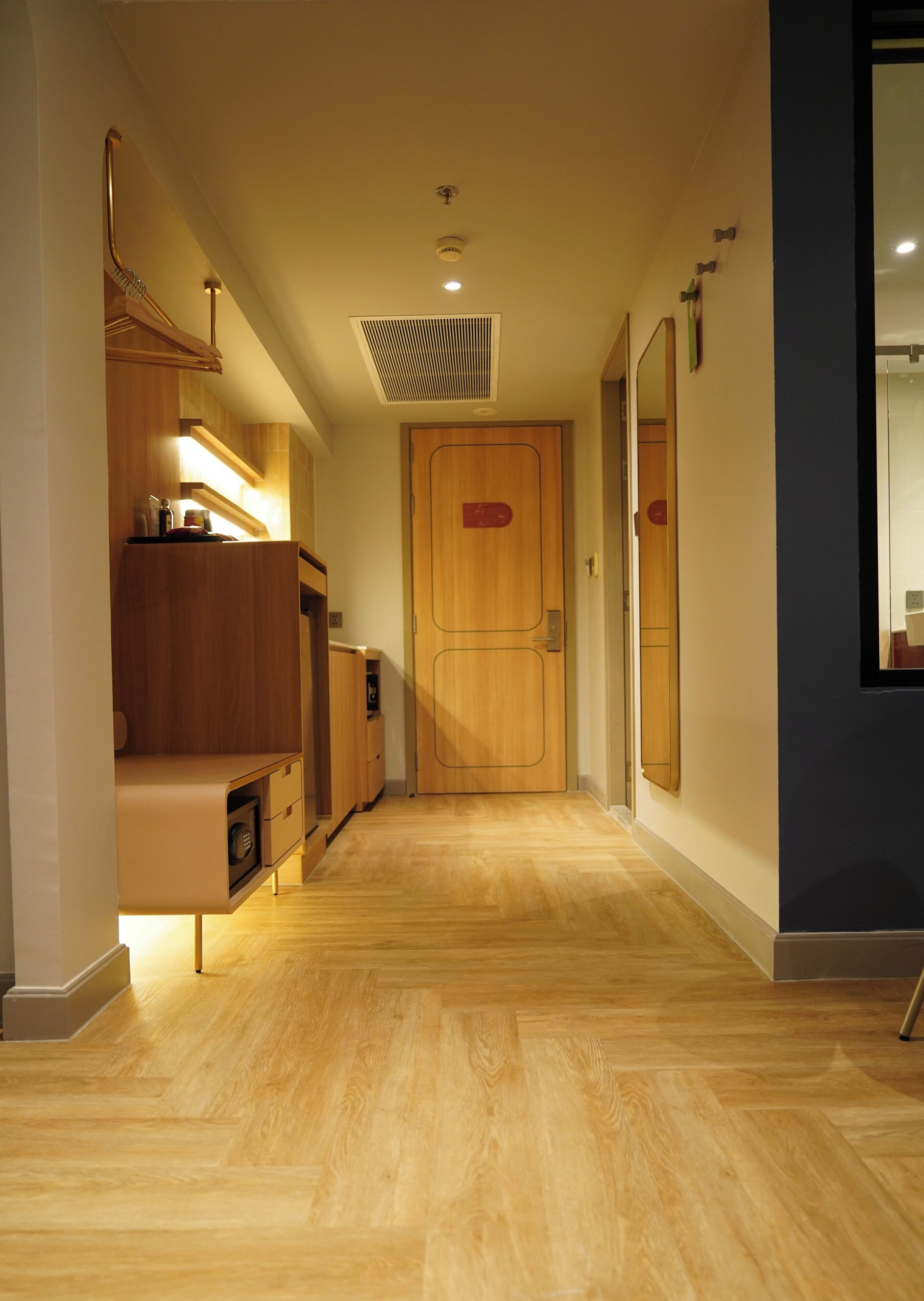 A hallway with wooden flooring, a wooden door, and a mirror on the right wall.