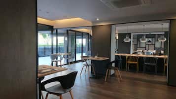 A modern dining area with wooden tables and chairs, a bar area with pendant lighting, and large windows providing natural light.