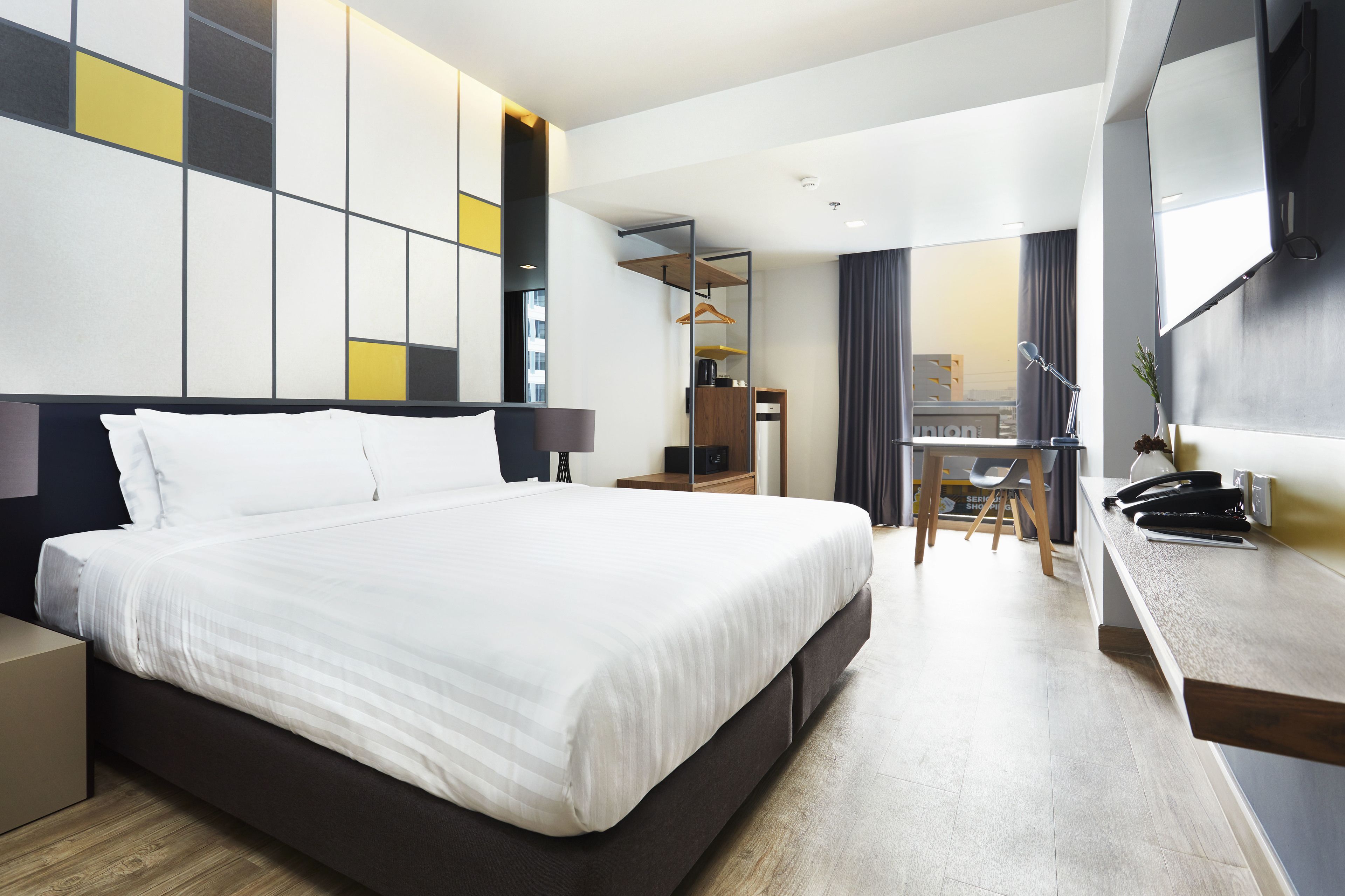A modern hotel room with a large bed, a desk with a chair, a wardrobe, and a TV.