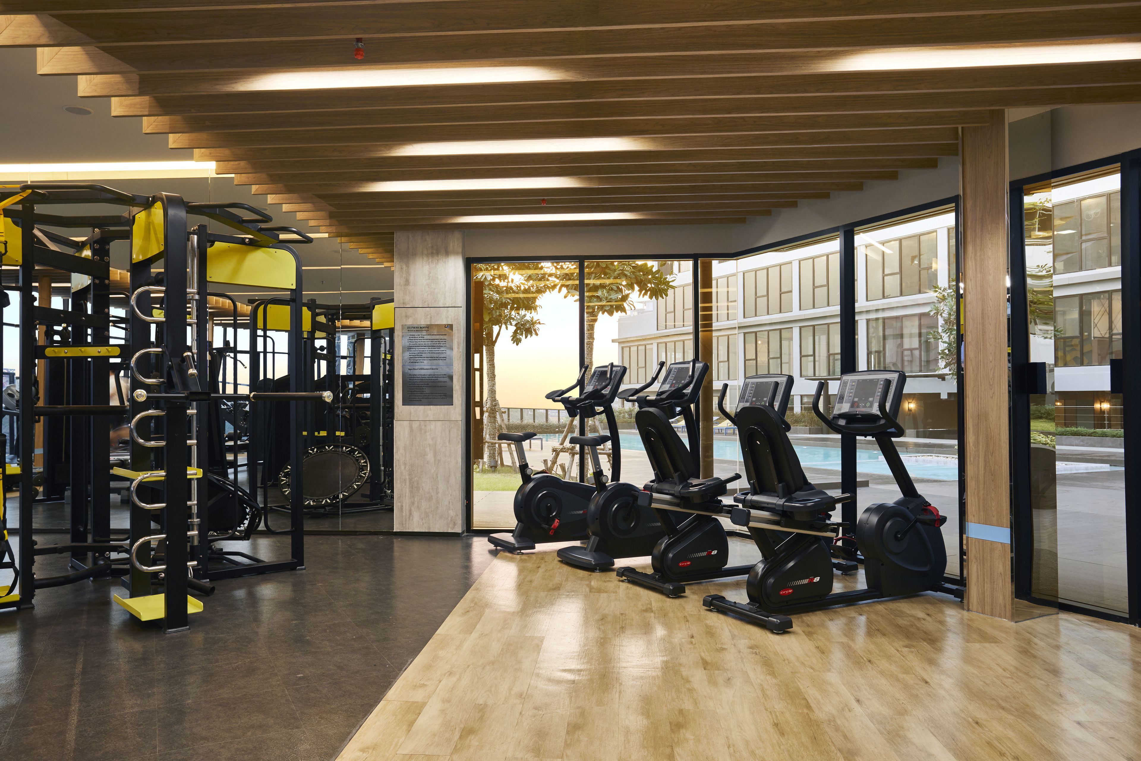 Un gimnasio con equipo de ejercicio, ventanas amplias y vistas a un edificio cercano.