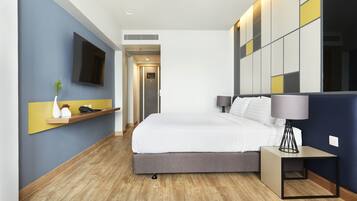 Una habitación de hotel moderna con una cama grande, una mesita de noche con lámpara, un televisor montado y un pequeño estante con objetos decorativos.